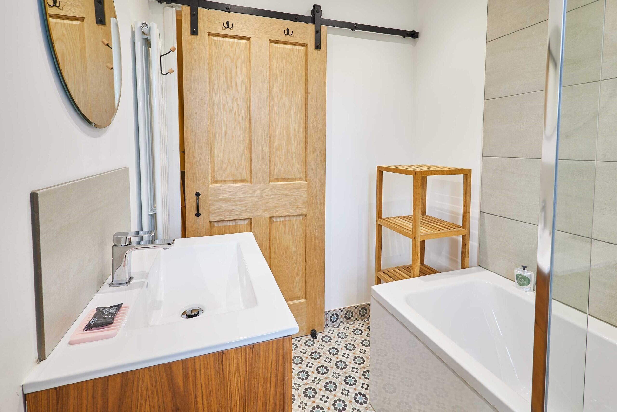 A bathroom with a wooden door, a white sink, a bathtub, and a shelf unit.