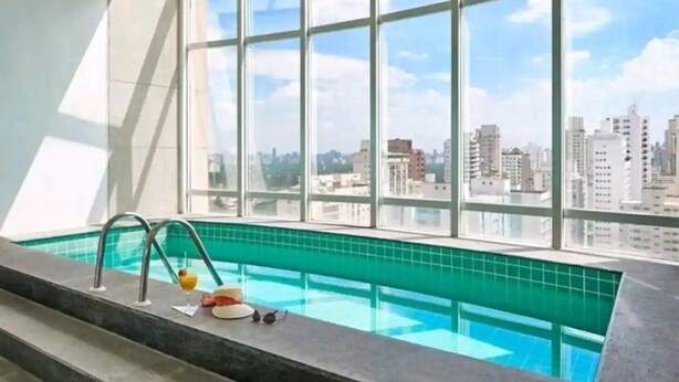 A modern indoor swimming pool with a city view, large windows, and a clear sky. - Loft-In Jardins Ibirapuera X (Paraíso São Paulo)