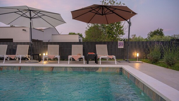 A swimming pool with lounge chairs and umbrellas, surrounded by a fence and a building in the background. - Poolside Holiday House (Labin)