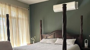 Ein Bett mit Baldachin, ein hölzerner Kopfteil, ein beiger Sessel und eine weiße Klimaanlage an der Wand.