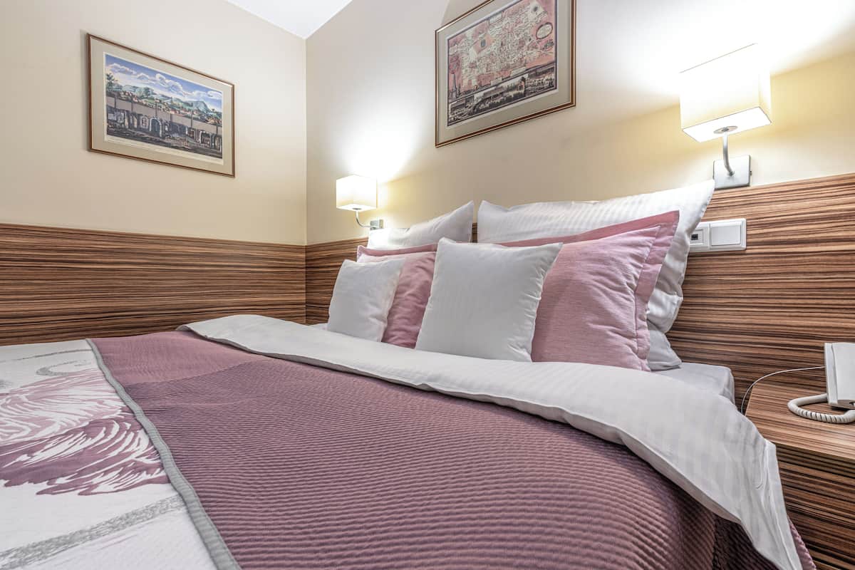 A neatly made bed with a pink and white bedspread, two wall-mounted bedside lamps, and two framed pictures on the wall.