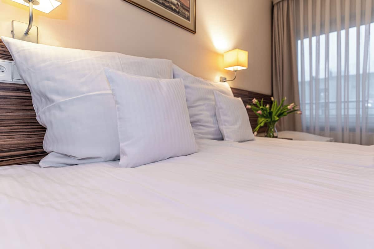 A hotel room with a neatly made bed, a wall-mounted lamp, a framed picture, and a window with curtains.