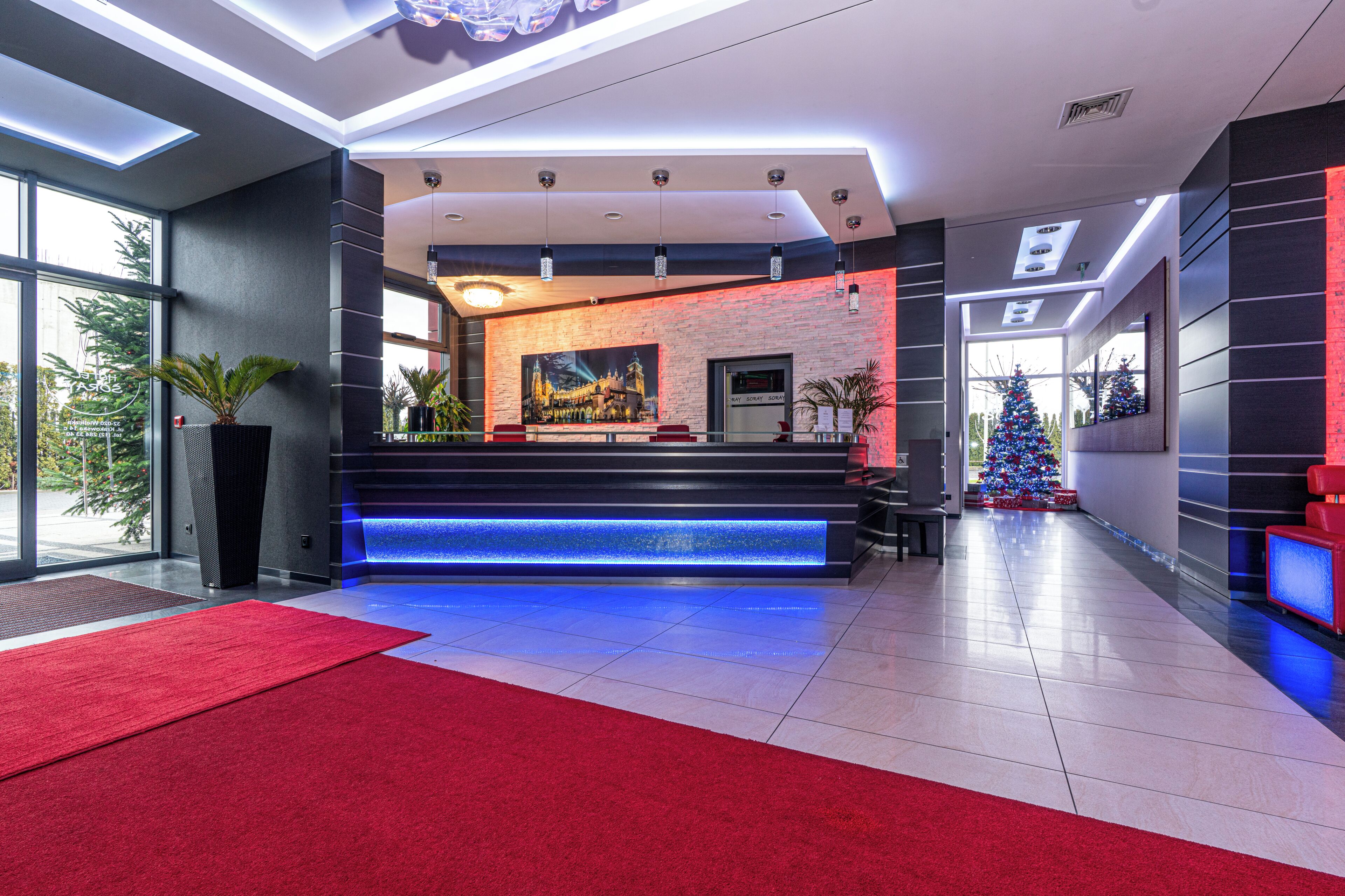 A modern hotel lobby with a reception desk, a large painting, a Christmas tree, and a red carpet.