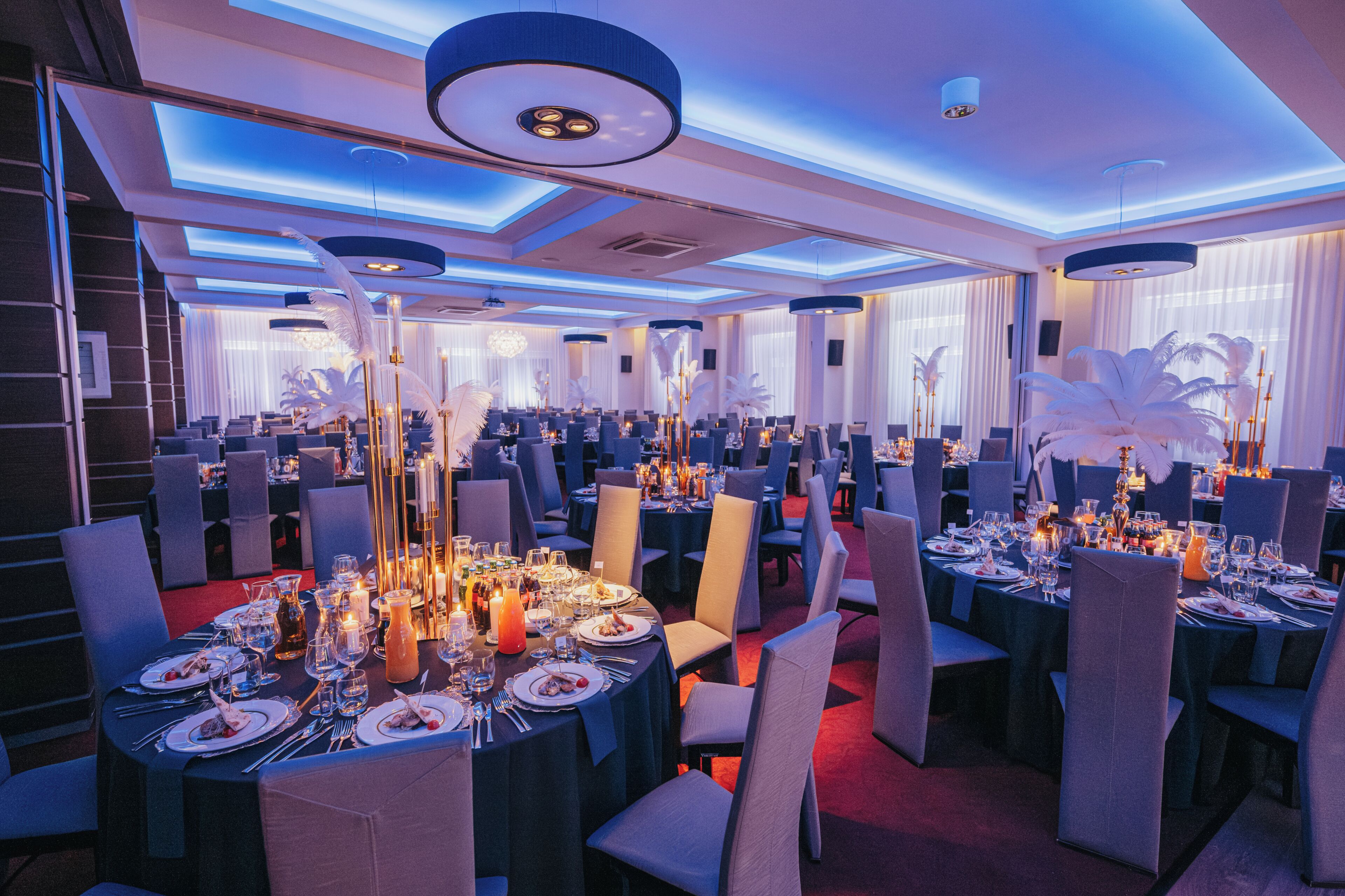 A banquet hall with round tables set for dining, featuring candle centerpieces and a modern ceiling design.