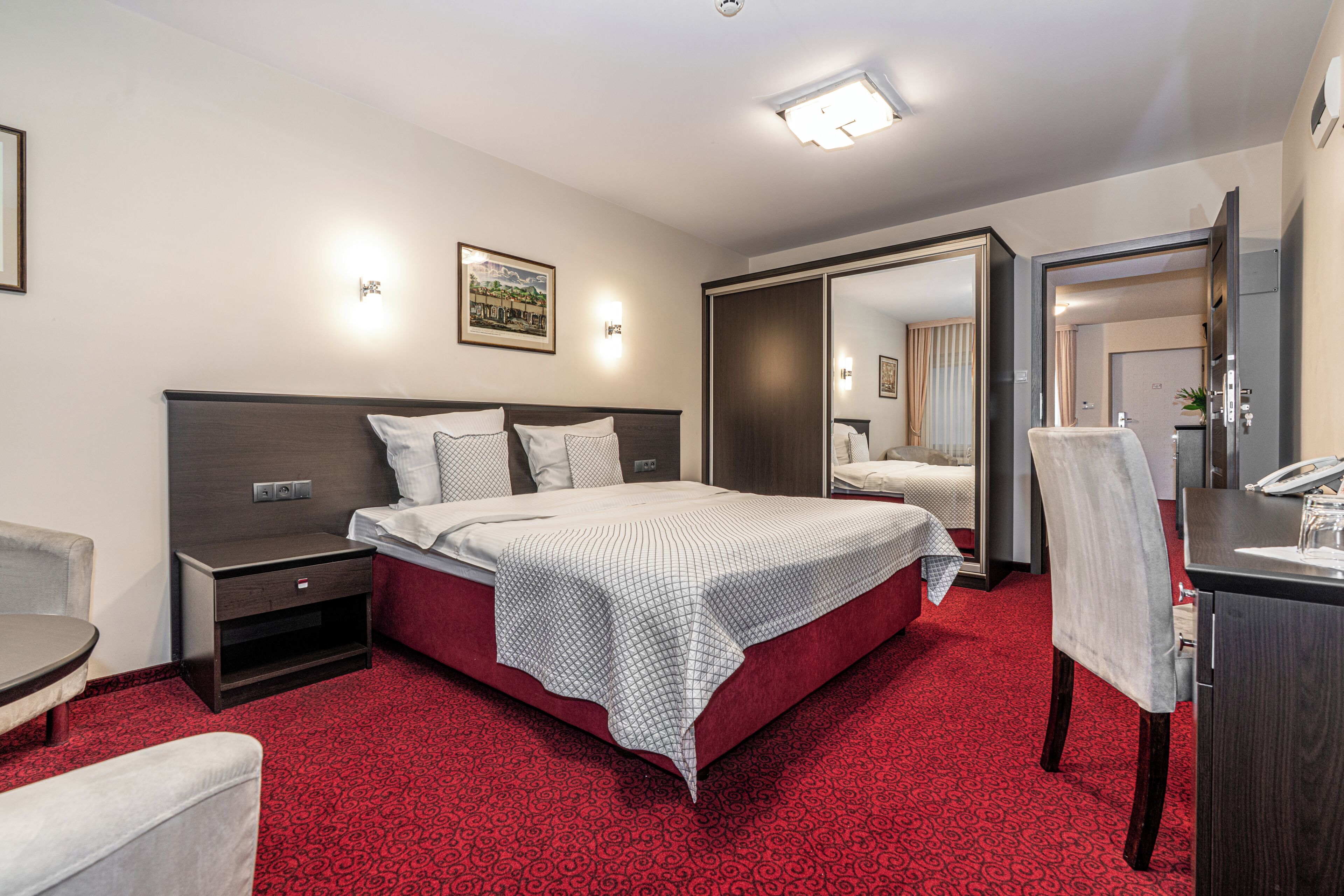 Quarto de hotel com cama, mesa de cabeceira, cadeira, escrivaninha e espelho.