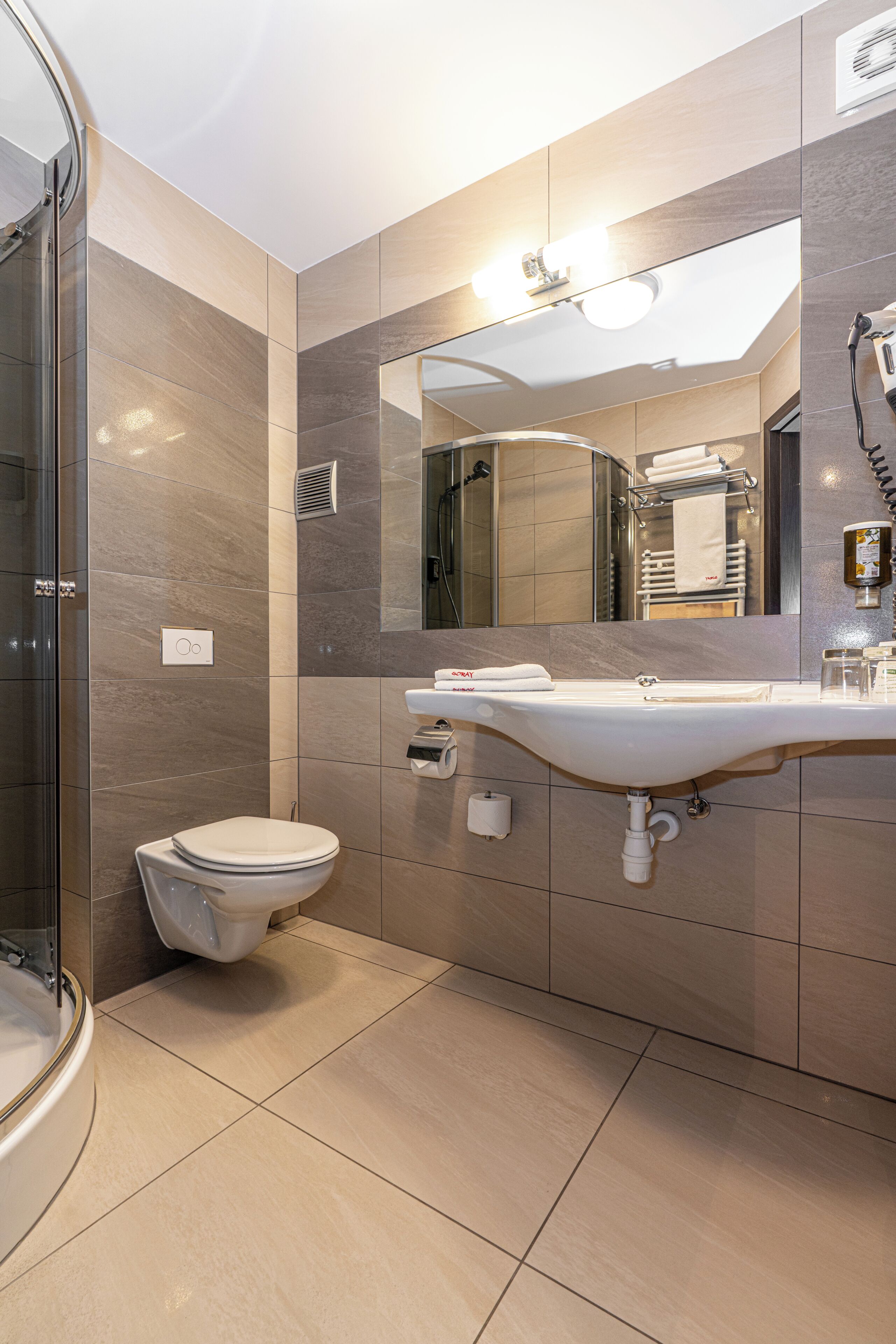 Une salle de bain moderne avec une douche à parois de verre, des toilettes blanches et un grand miroir au-dessus d’un lavabo.