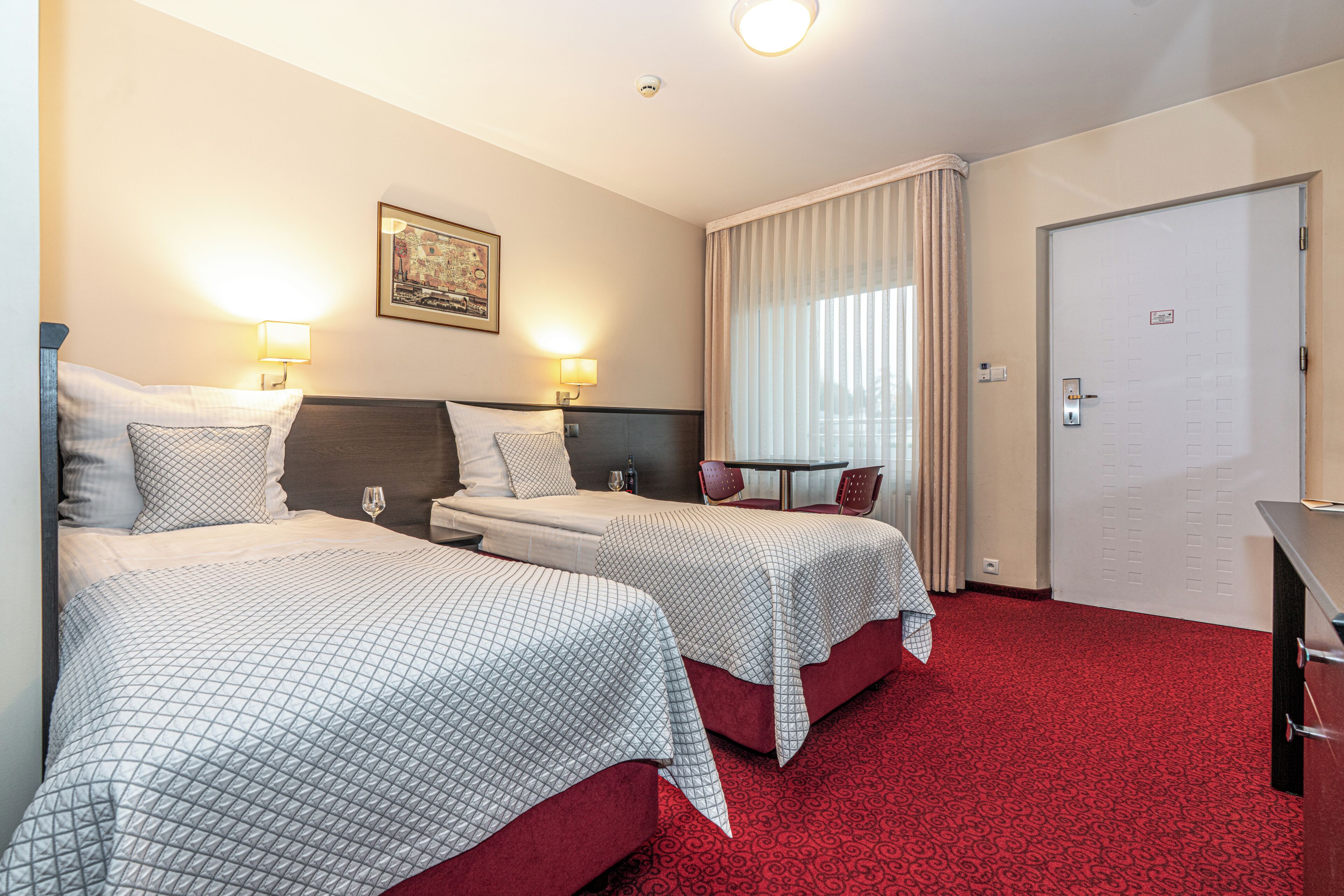 Quarto de hotel com duas camas, um tapete vermelho, um quadro na parede e uma porta com fechadura.