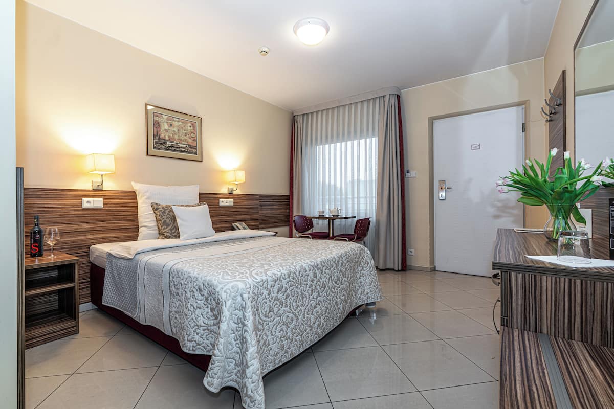 A hotel room with a bed, bedside lamps, a desk with a vase, and a door.