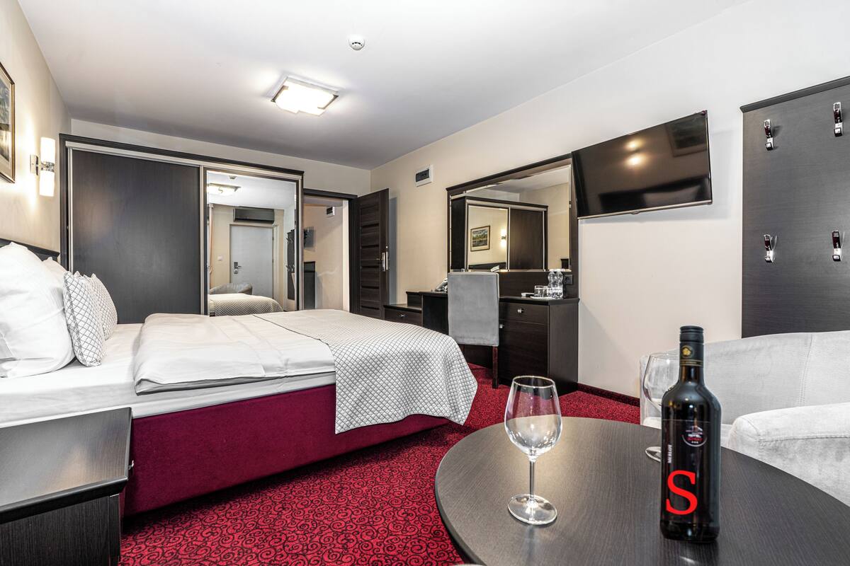 A hotel room with a bed, a desk, a table with a bottle and glass, a TV, and a mirror.