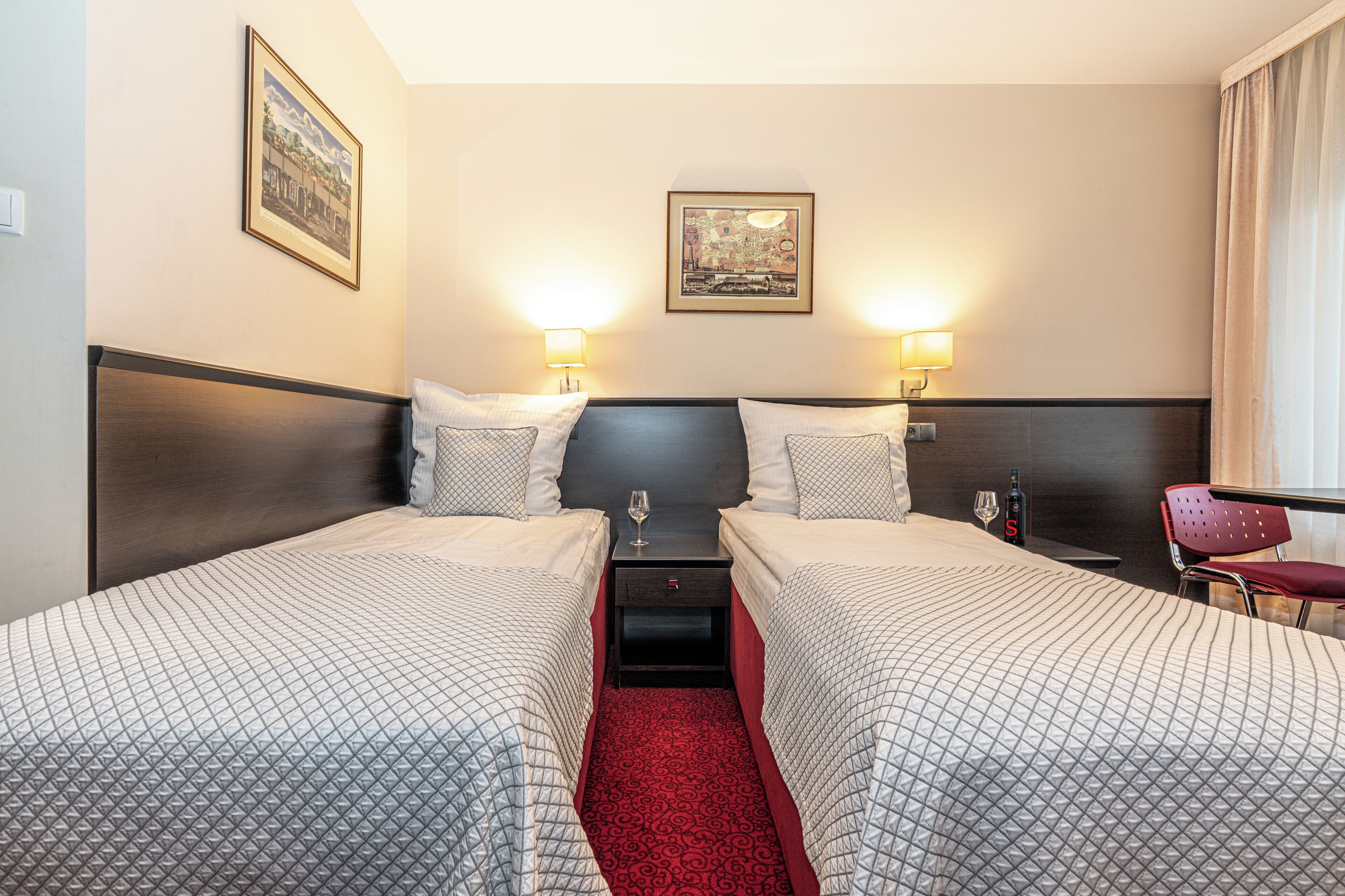 A hotel room with two beds, a red carpet, a nightstand with a wine glass, and a chair.