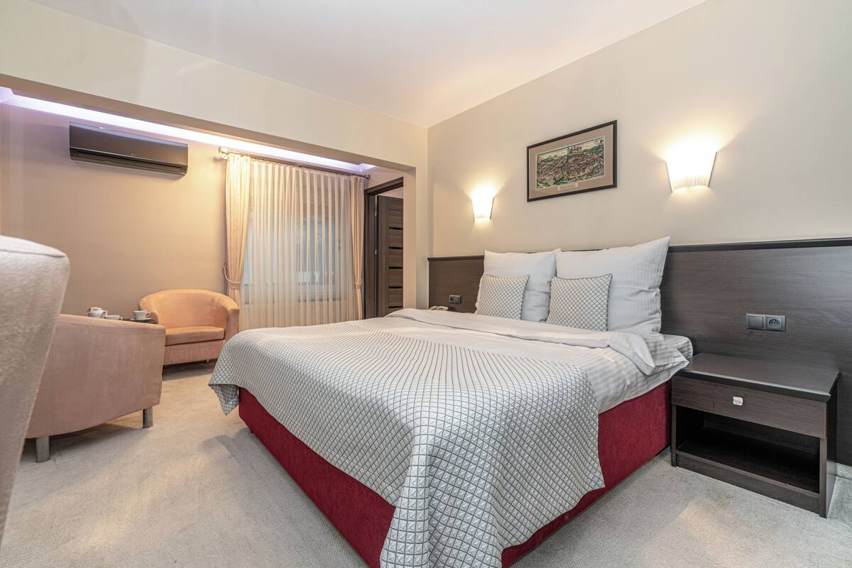 A hotel room with a bed, a chair, a bedside table, an air conditioning unit, a wall-mounted light, and a framed picture.