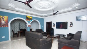 Un salon avec des canapés sombres, une table basse et un téléviseur. On y trouve un ventilateur de plafond, un tableau au mur et une porte au fond.