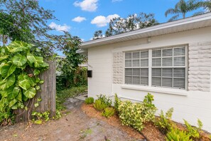 Uma casa branca com fachada de pedra, uma janela com persianas e um jardim com diversas plantas e cerca de madeira.