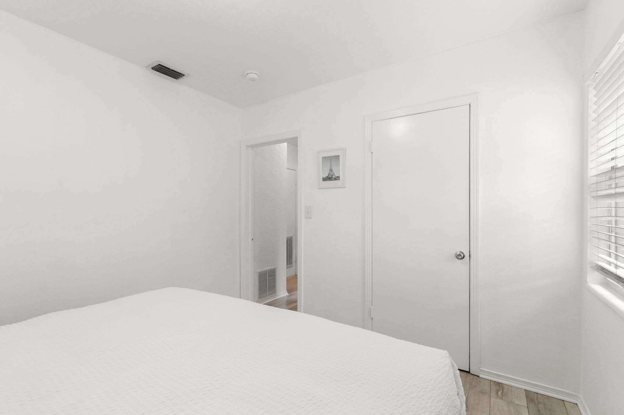 Ein helles Schlafzimmer mit Bett, einer Tür und einem Fenster mit Jalousien.