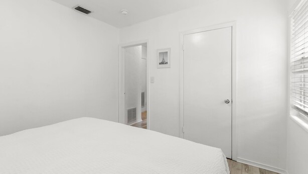 A white bedroom with a bed, a door, and a window with blinds. - Bayside Escape | Cozy Hideaway Near Bayshore (Tampa)