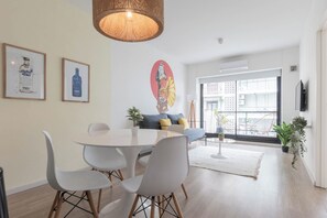 A modern living room with a round dining table, white chairs, a sofa with yellow cushions, a large window, and a wall-mounted TV. - MOT Zapiola (Buenos Aires)