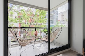 A balcony with wicker chairs and a table, a large window, and a view of buildings and trees.
