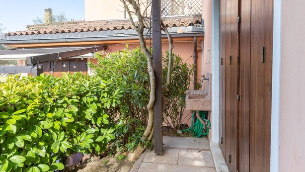 A narrow pathway leading to a wooden door, flanked by lush green foliage and a tiled roof in the background. - Tina Home Lake View (Garda)