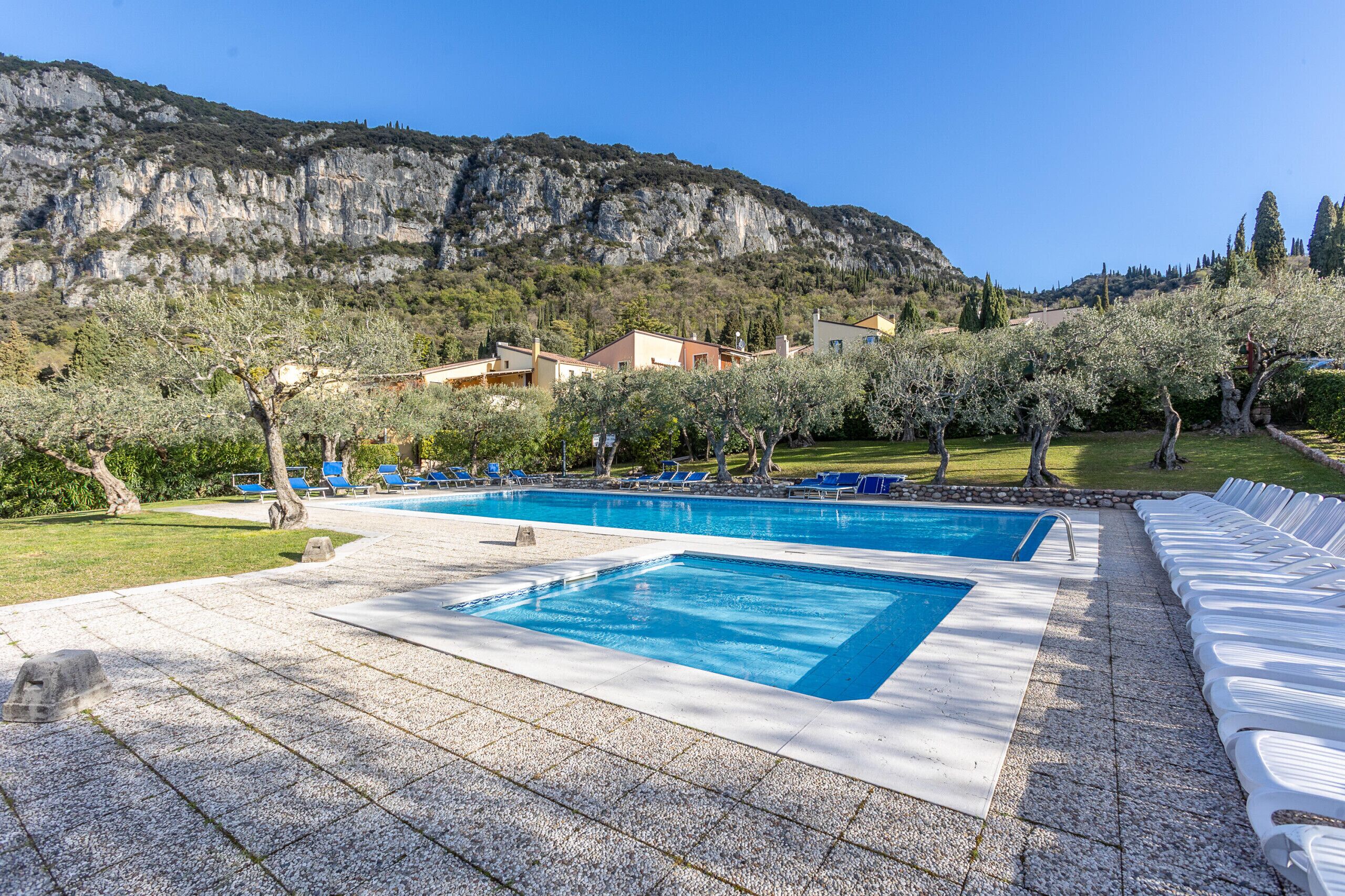 Area piscina con due piscine rettangolari, circondata da lettini e alberi, con montagne e edifici sullo sfondo.