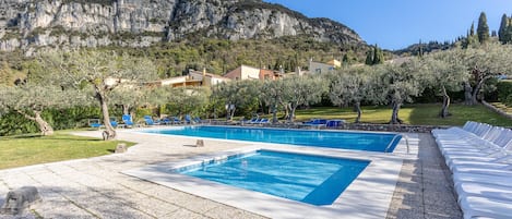 A pool area with two rectangular pools, surrounded by lounge chairs and trees, set against a backdrop of mountains and buildings.