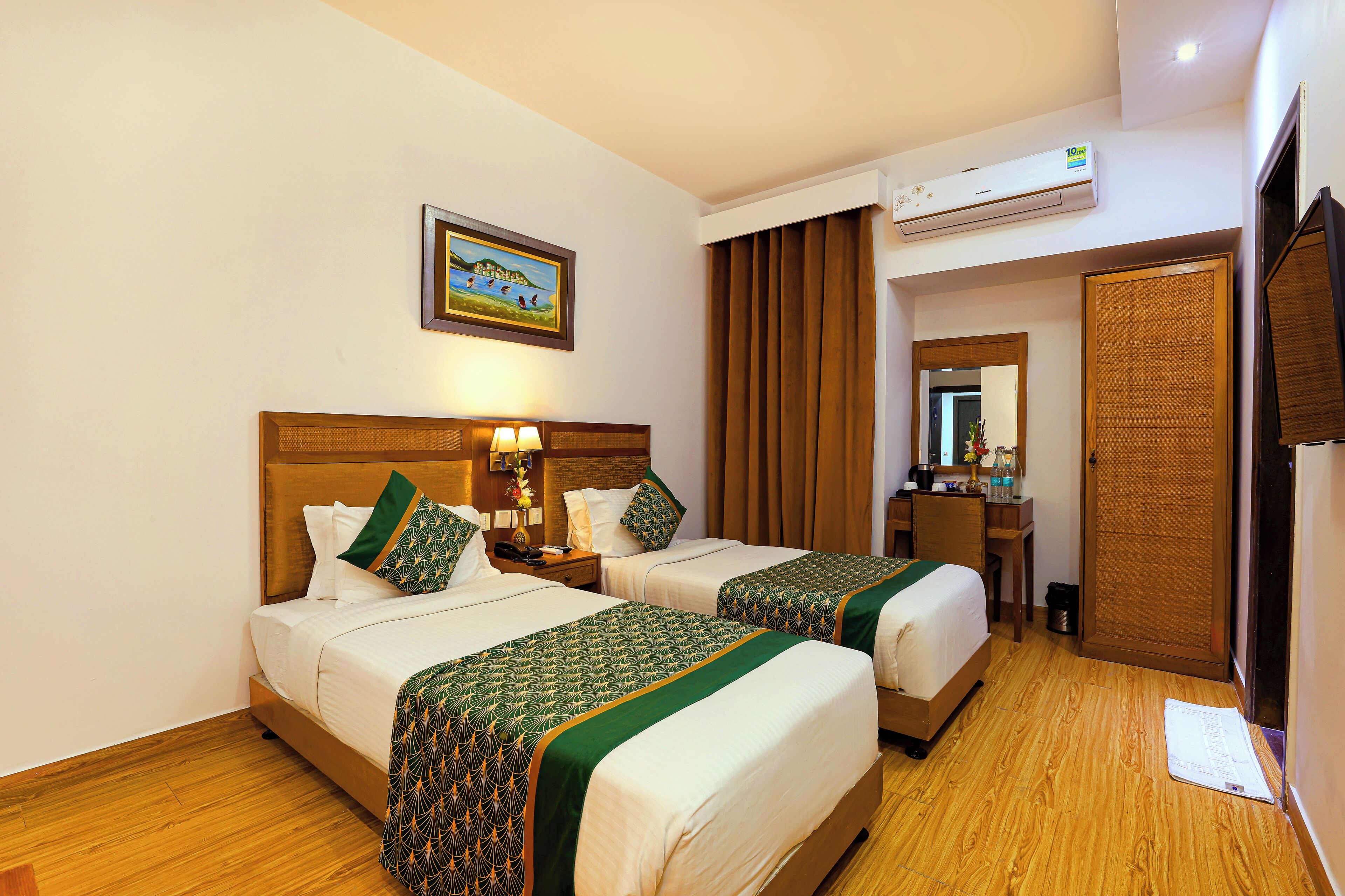 A hotel room with two beds, wooden flooring, an air conditioning unit, a desk with a mirror, and a painting on the wall.