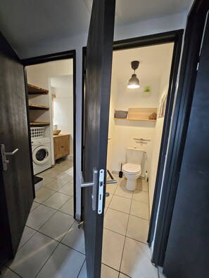Eine geöffnete Tür zu einem Waschraum mit Waschmaschine und Regalen, einer Toilette mit Bidet sowie einer Pendelleuchte.