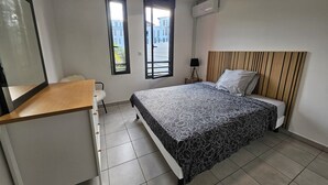 Un dormitorio con cama, escritorio, dos ventanas, aire acondicionado y vista a edificios.