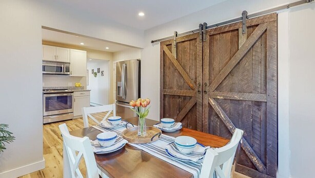 A dining area with a wooden table set for four, white chairs, and a rustic barn door in the background. - Enchanting 3BR Farmhouse Retreat  Nature Getaway (Gorham)