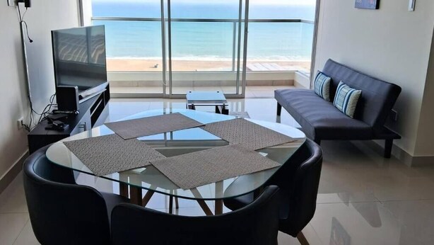 A modern living room with a glass table, black chairs, a sofa with patterned cushions, a flat-screen TV, and a view of the ocean. - Beautiful apartment with balcony facing the sea (Lima)