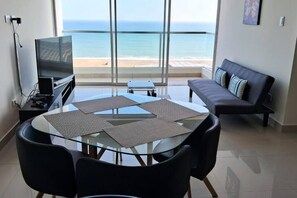 A modern living room with a glass table, black chairs, a sofa with patterned cushions, a flat-screen TV, and a view of the ocean. - Beautiful apartment with balcony facing the sea (Lima)