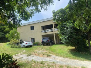 A two-story building with a balcony on the upper floor, a covered parking area, and a grassy lawn with a gravel path leading to the entrance. - 3-bedroom house in fabulous St Thomas with AC, WiFi. Enjoy your stay (St Thomas)