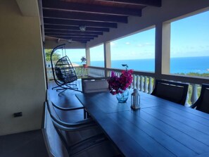 A balcony with a wooden table, chairs, a hanging chair, and a view of the ocean. - 3-bedroom house in fabulous St Thomas with AC, WiFi. Enjoy your stay (St Thomas)