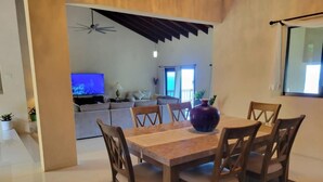 Un comedor con una mesa y sillas de madera, un jarrón morado y vista a una sala de estar con televisor y sofá.