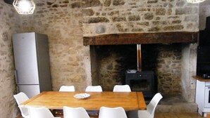 Een kamer met stenen muren, een houten tafel, witte stoelen, een open haard en een moderne keukenhoek.