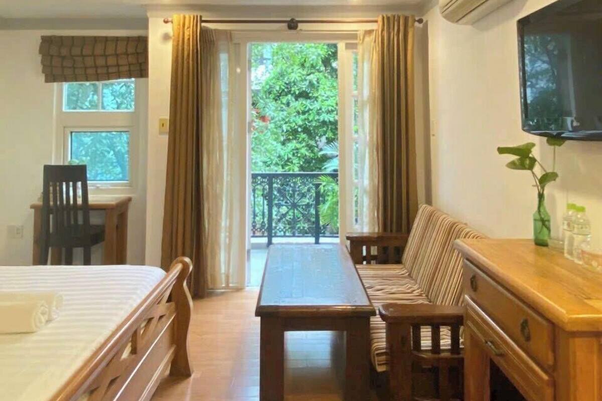 A room with a bed, a dining table, and a sofa. There is a window with curtains, an air conditioning unit, and a TV mounted on the wall.