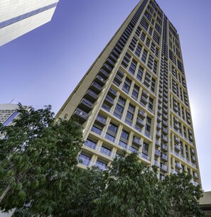 Een hoog, modern gebouw met een raster van ramen en balkons, omringd door weelderige groene bomen.