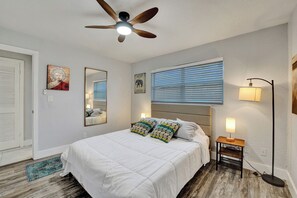Ein Schlafzimmer mit Bett, Deckenventilator, Nachttisch mit Lampe, Fenster mit Jalousien und Teppich.