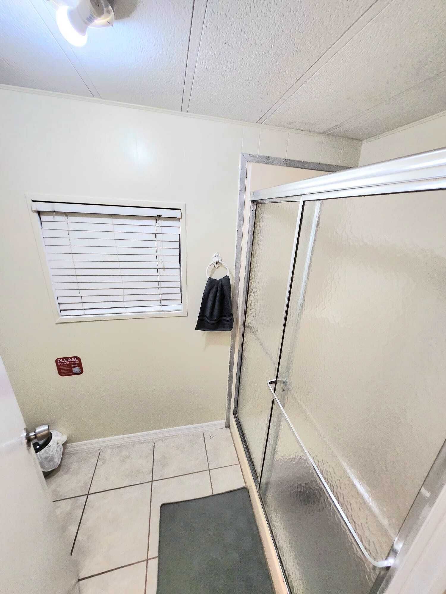 Un espacio de ducha pequeño y cerrado con puerta de vidrio, una ventana con persianas, y una toalla colgada en un gancho.