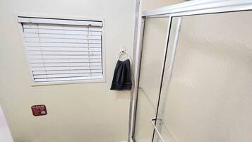 Un espacio de ducha pequeño y cerrado con puerta de vidrio, una ventana con persianas, y una toalla colgada en un gancho.