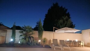 Un espace extérieur avec une piscine, des chaises longues et une table placée sous un parasol, le tout entouré de plantes et d’arbres.