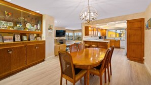 A spacious dining area with a wooden table and chairs, a chandelier, and a kitchen in the background. - 25% Off Sept 10-12, Walk 2 Beach, Private Jacuzzi! (Oxnard)