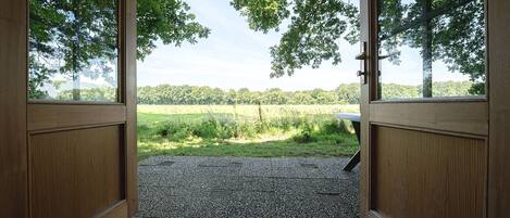 Een open houten deur die uitkomt op een grindpad, met uitzicht op een grasveld en bomen.