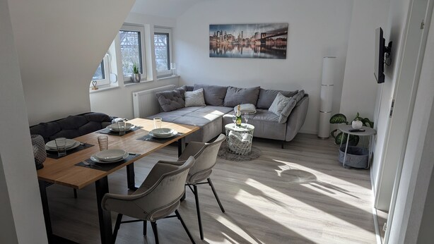 A modern dining area with a wooden table set for two, a grey sofa, and a cityscape painting on the wall. - Charming vacation apartment in the heart of the northern Black Forest (Haiterbach)
