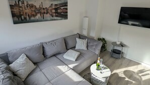 A modern living room with a grey sectional sofa, a cityscape painting, a flat-screen TV, and a small round coffee table with a bottle and glass. - Charming vacation apartment in the heart of the northern Black Forest (Haiterbach)