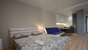 Une chambre moderne avec un grand lit, un canapé bleu et, au fond, un coin cuisine.