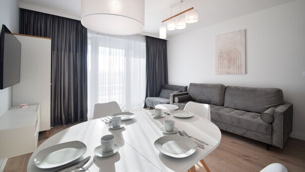 A modern, well-lit dining area with a round table set for four, a grey sofa, and a flat-screen TV mounted on the wall. - RentPlanet - Apartament Marina (Trzebiatow)