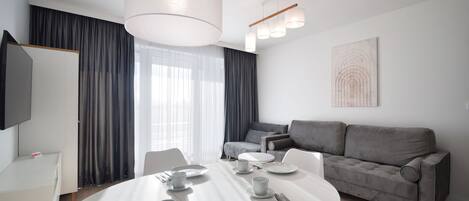 Een moderne, goed verlichte eetruimte met een ronde tafel gedekt voor vier personen, een grijze bank en een flatscreen televisie aan de muur.