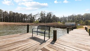 Un muelle de madera con un banco, una casa pequeña al fondo y un área boscosa al otro lado del agua.