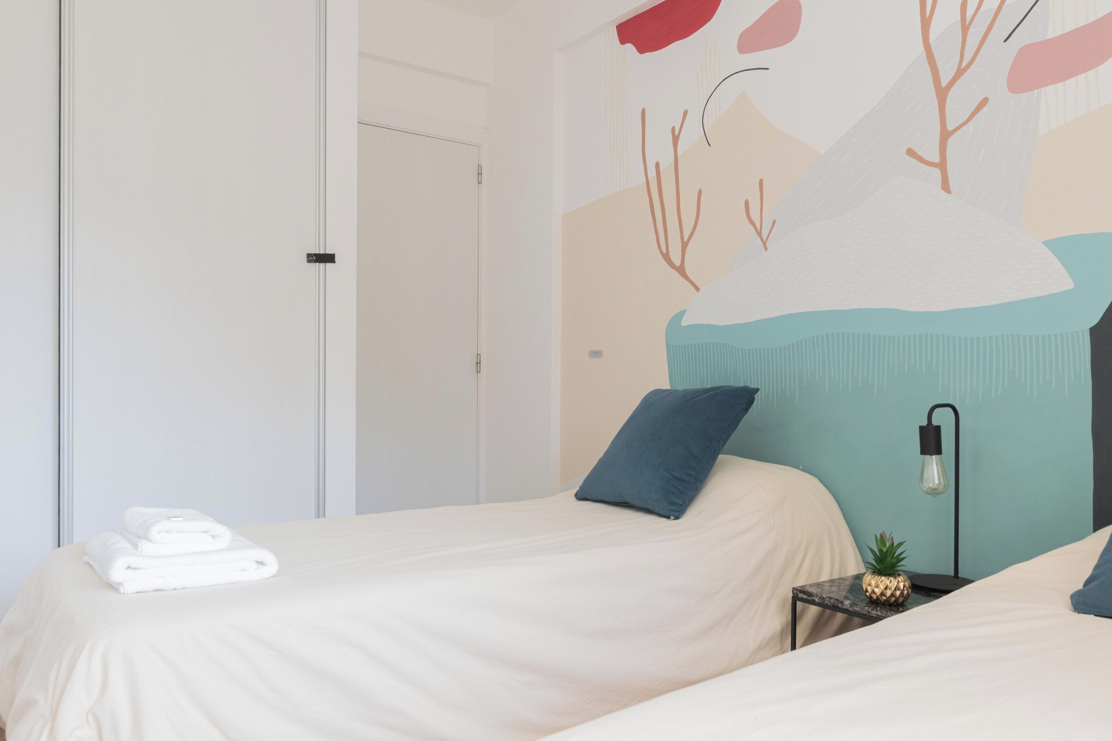 A hotel room with two beds, a door, a wall with abstract art, a nightstand with a lamp, and a pineapple on the nightstand.
