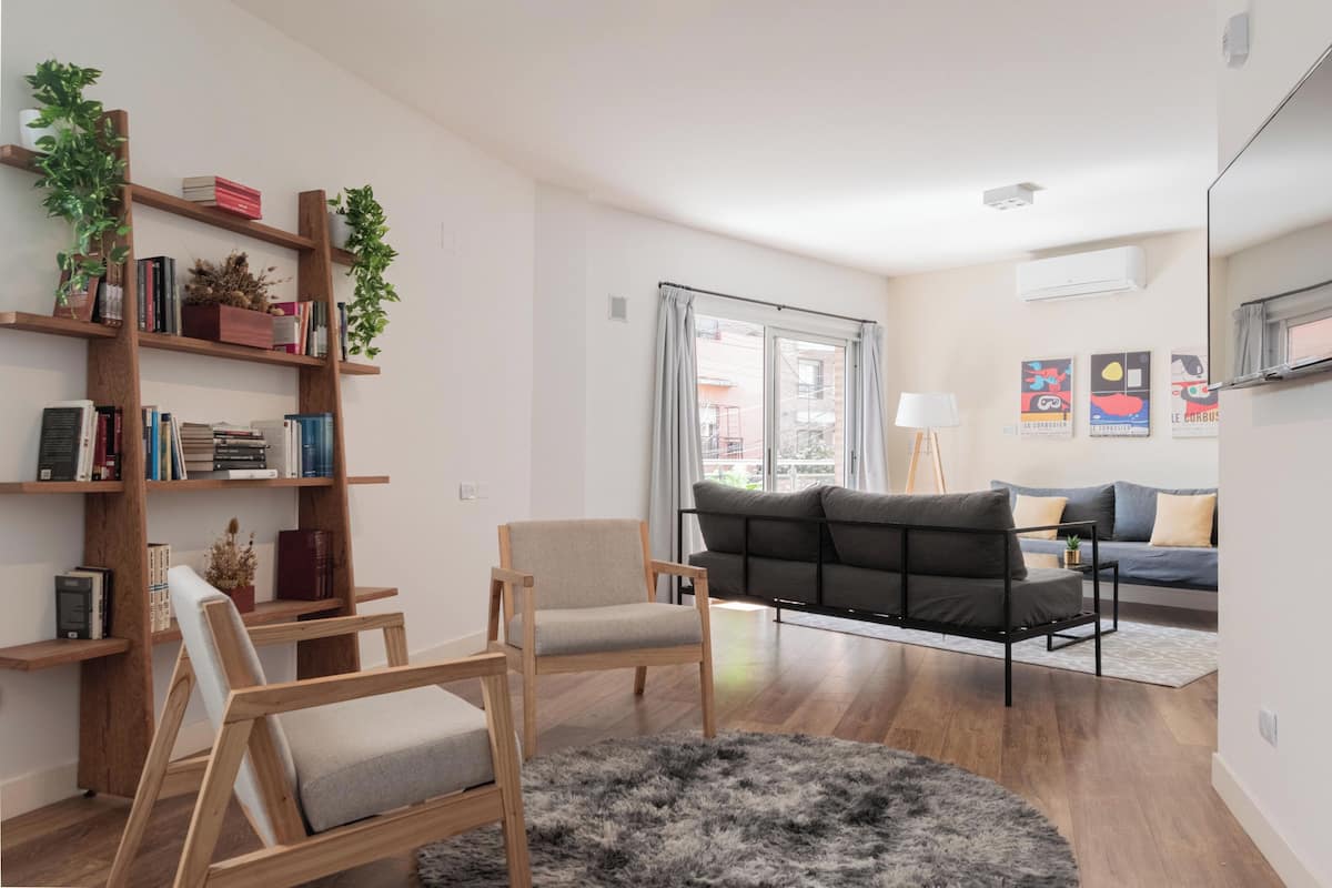 A modern living room with a wooden bookshelf, a grey armchair, a black sofa, and a large window with curtains.